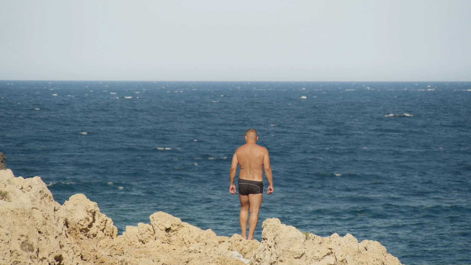 A figure faces the sea from the edge of a rocky cliff.