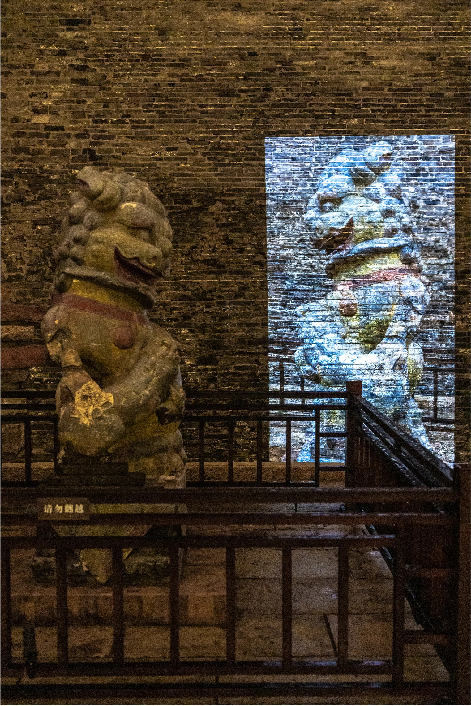 A stone lion and its projected counterpart stand against a brick wall at night.