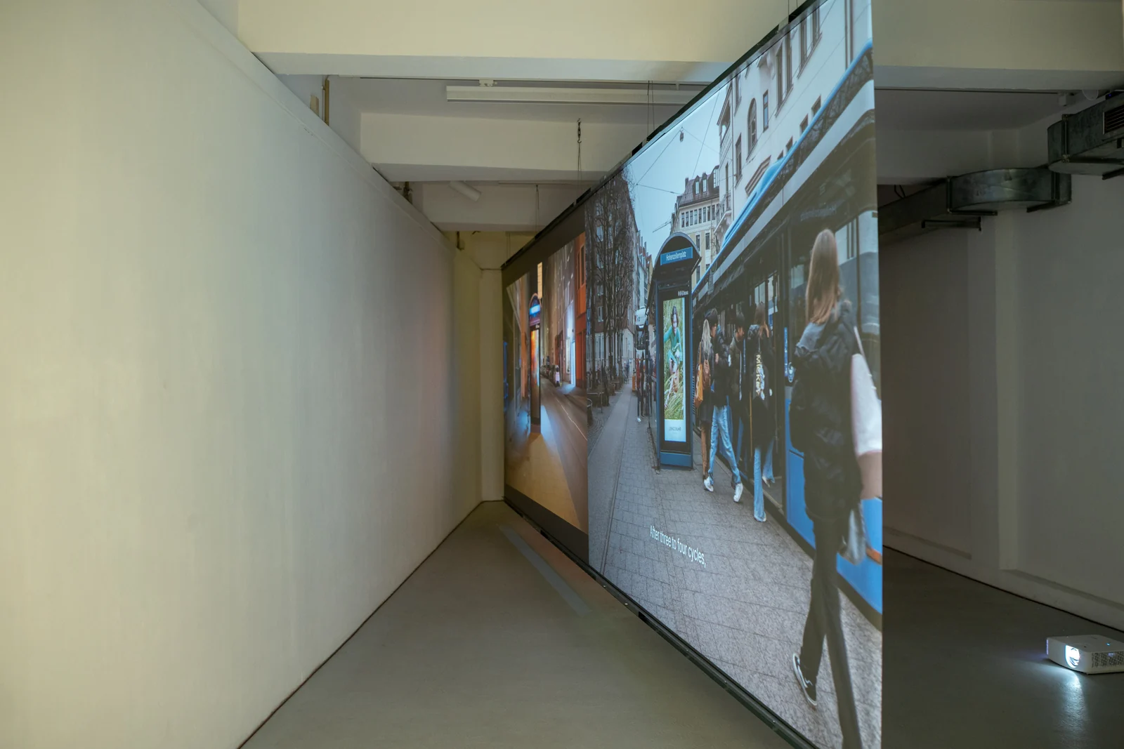 Oblique view along a large suspended projection screen, with a white gallery wall on the left.