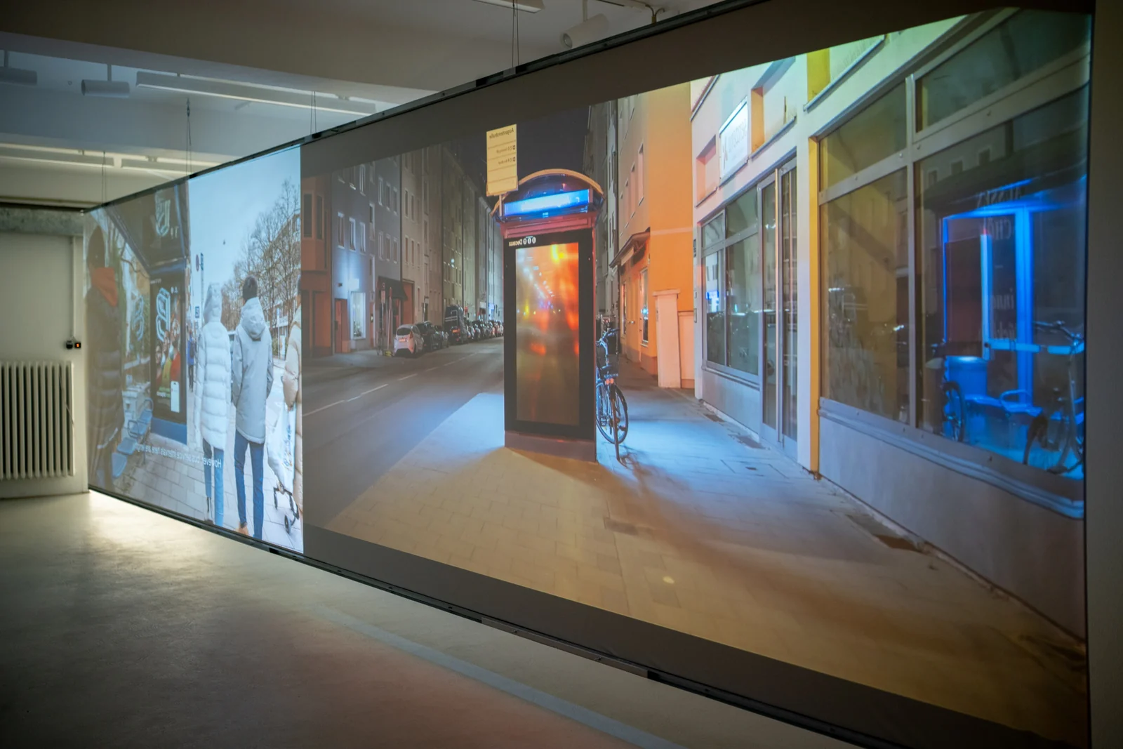 Two projected street scenes fill a suspended screen in a gallery; an illuminated billboard appears on the right.