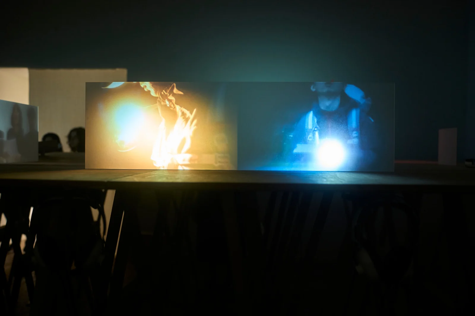A dual-channel projection in a dark gallery, with warm yellow and cool blue beams of light.