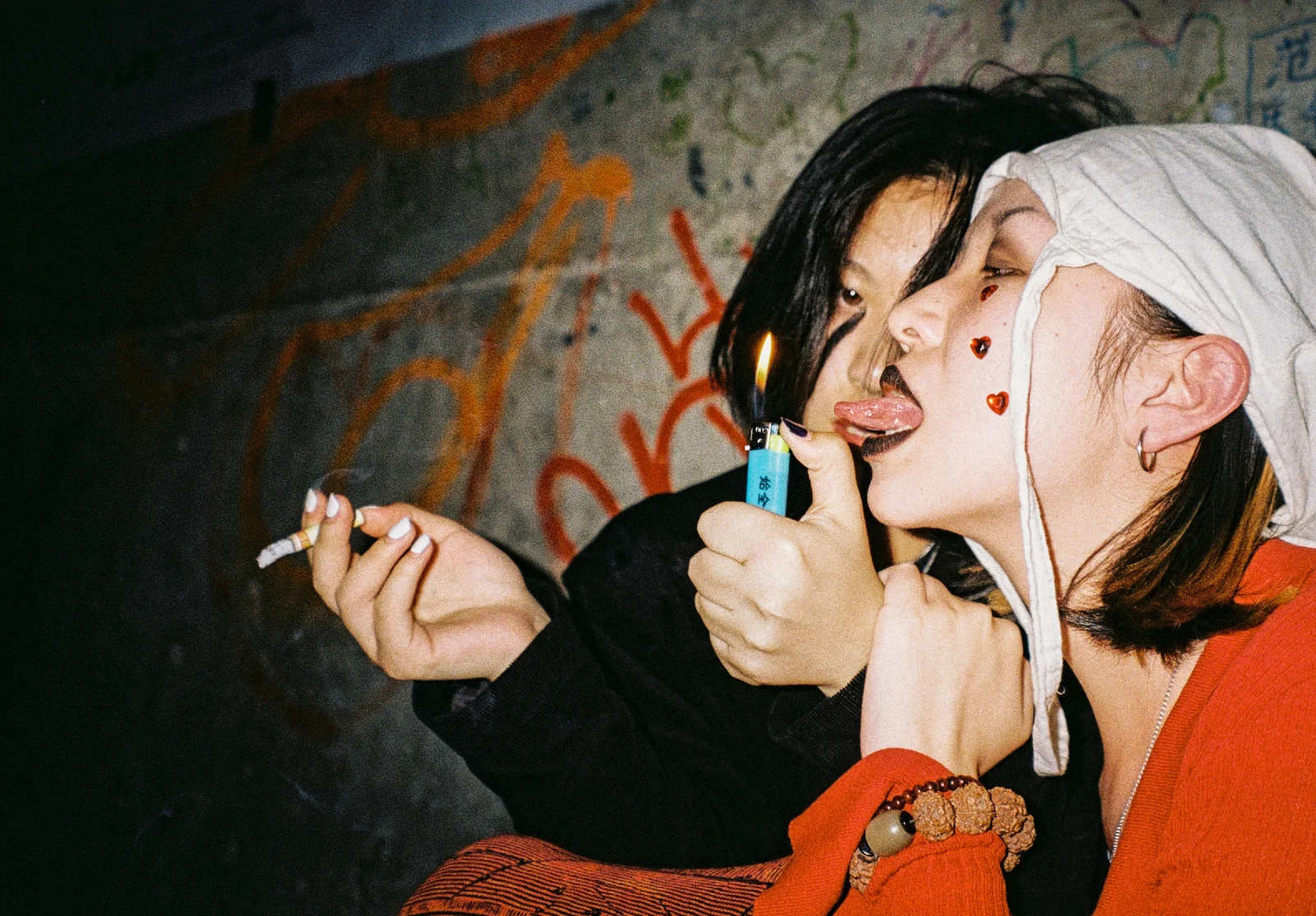 Two young people stand closely together beside a graffiti-covered wall, lit by direct flash.
