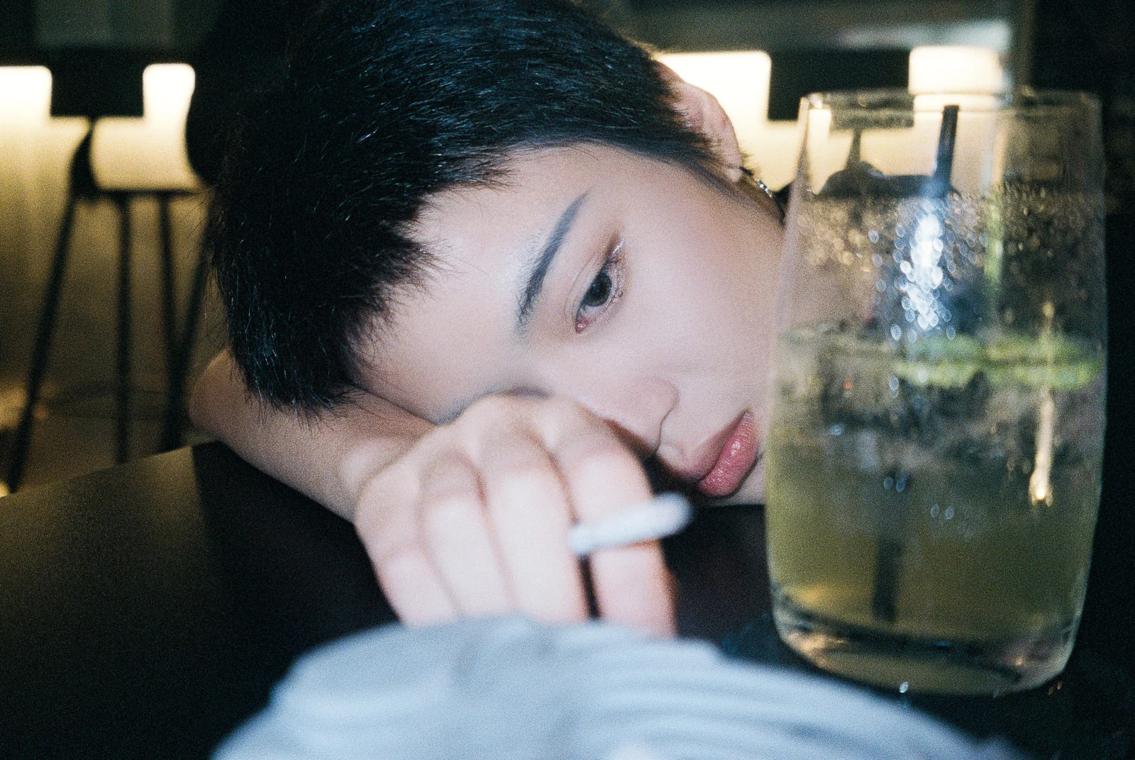 A person rests their head on an arm at a table beside a glass.