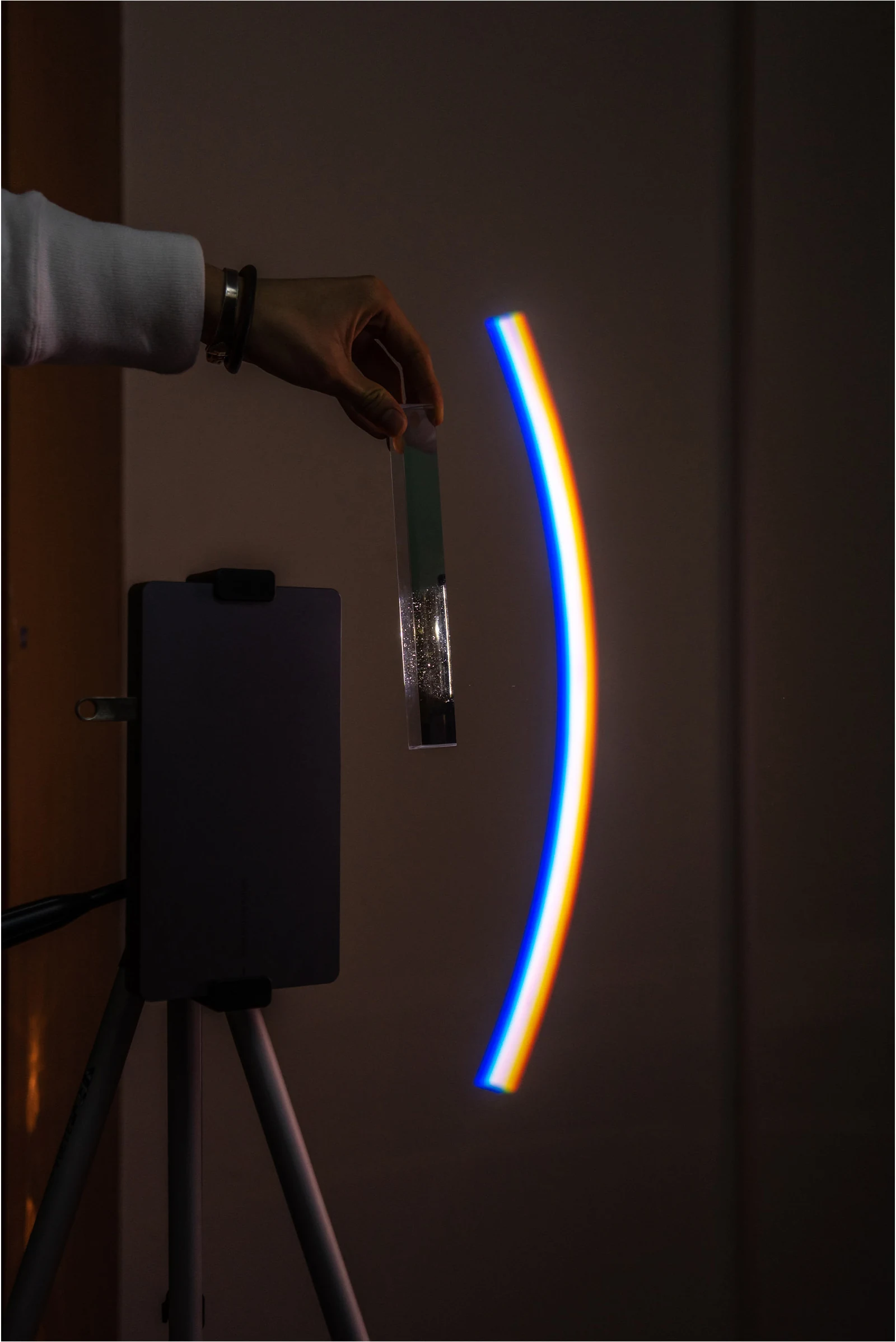 A hand holds a triangular prism beside a rainbow projected onto a wall.