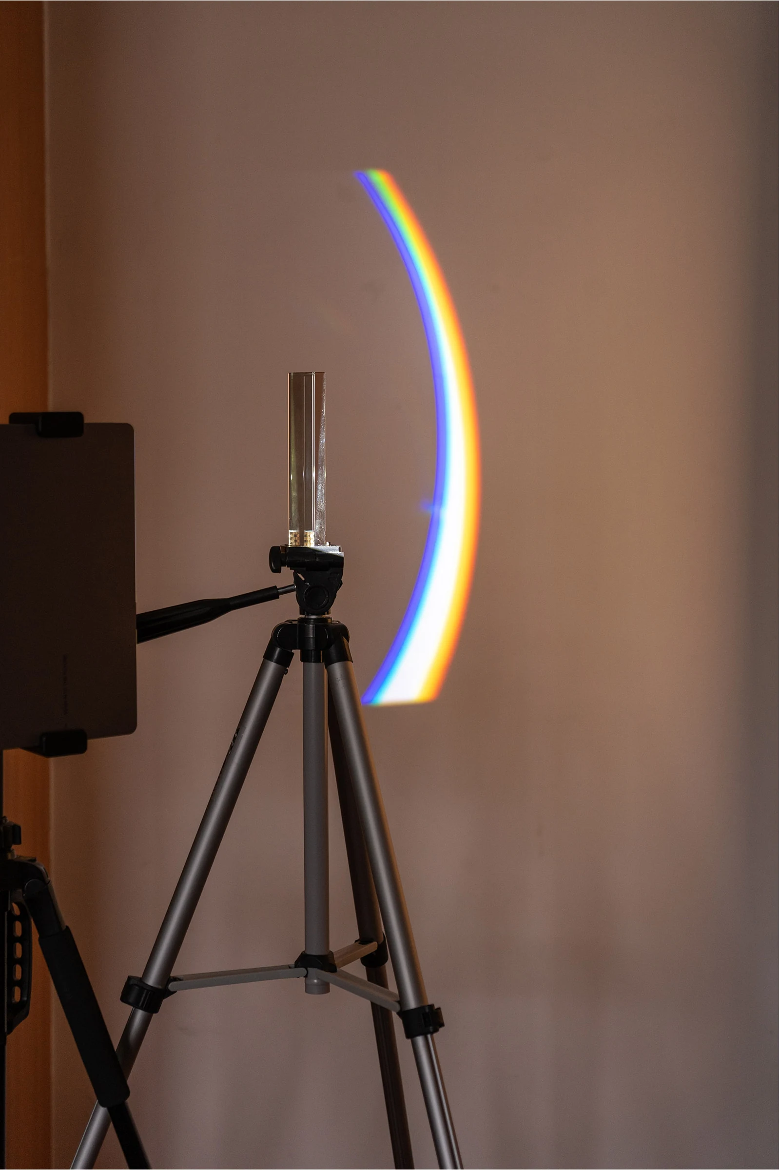 A triangular prism on a tripod refracts projected light into a curved rainbow.